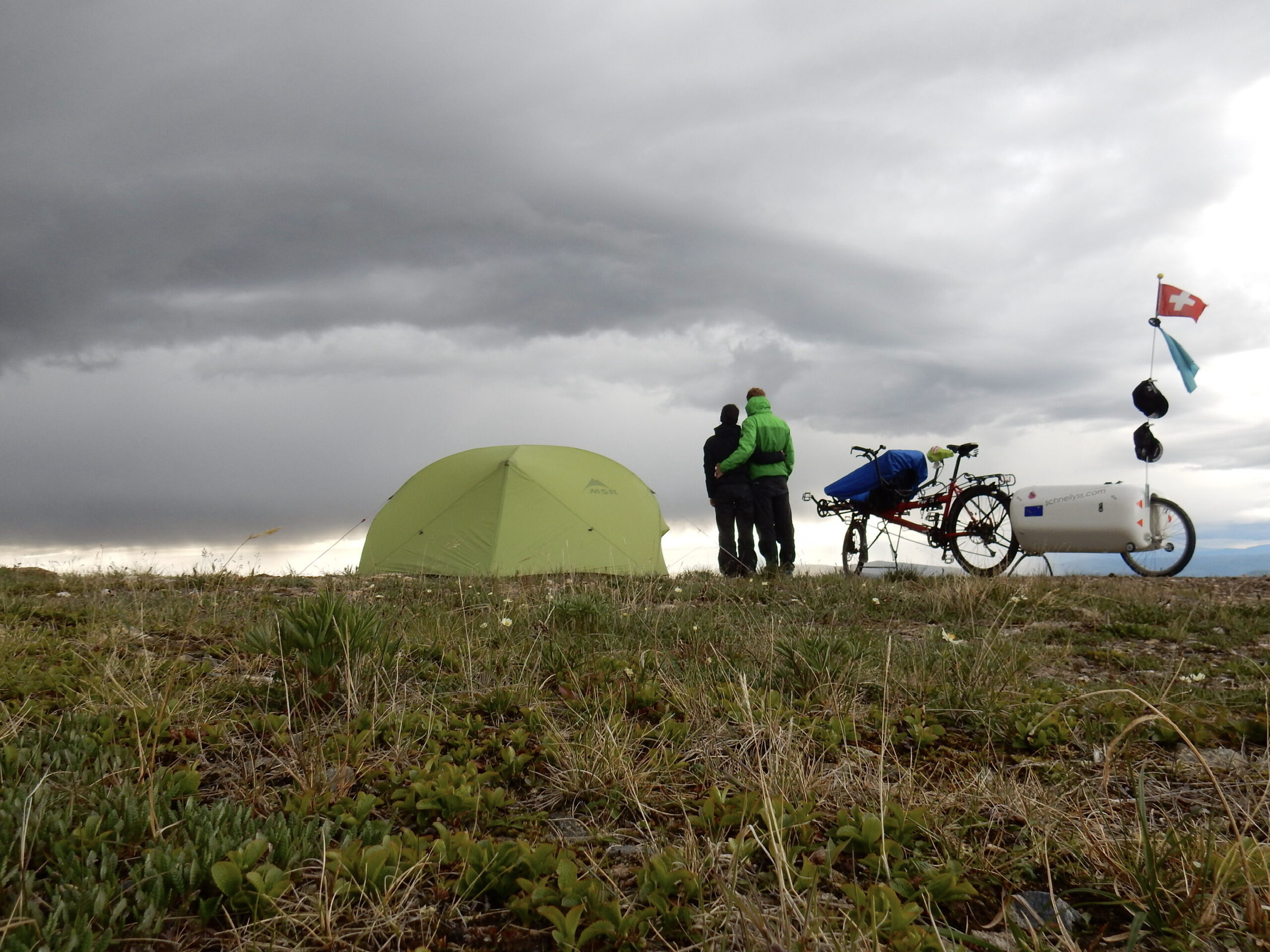 Deux voyageurs près d’une tente, d’un tandem et d’une remorque sous un ciel nuageux